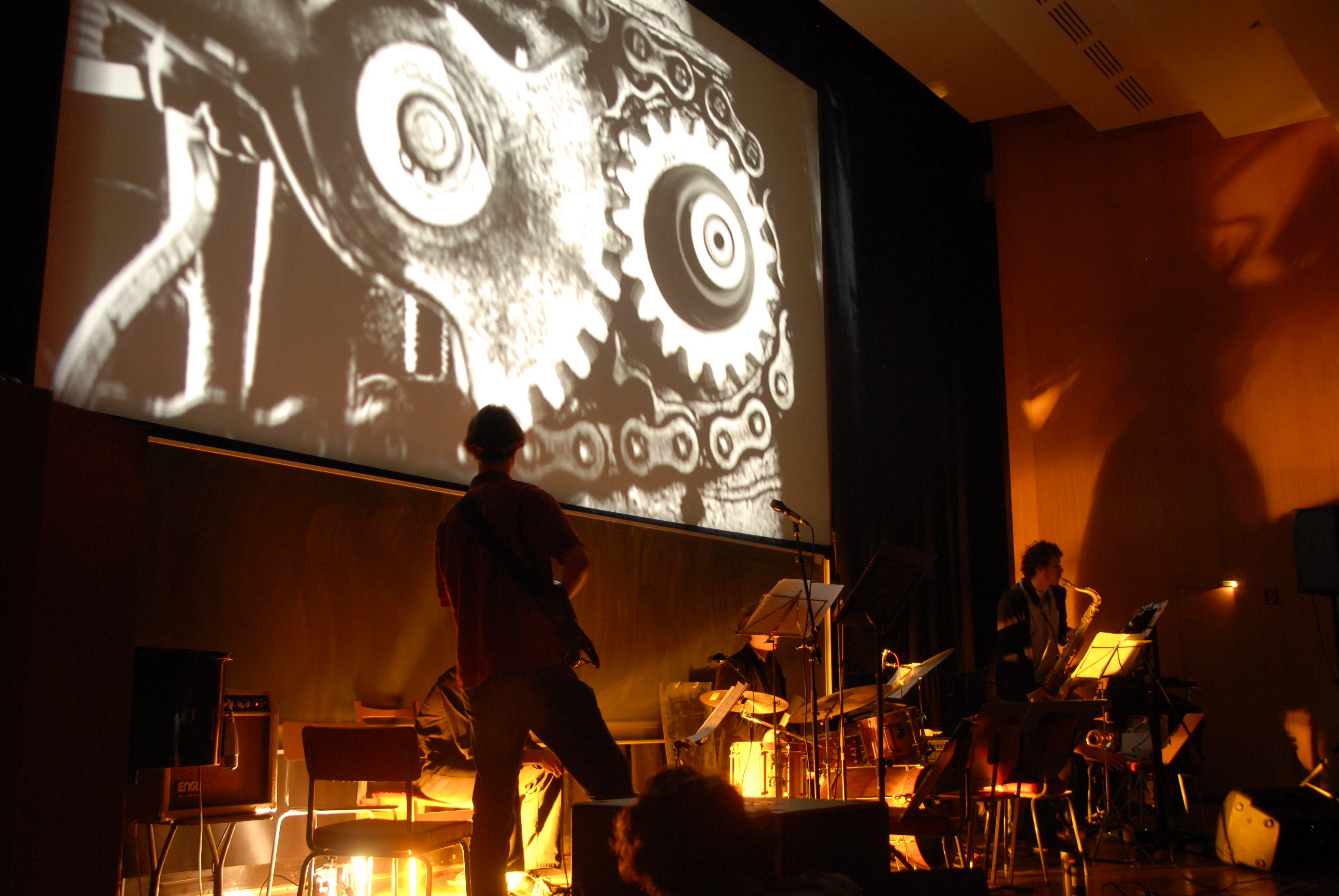 Three musicians with guitar, drums and saxophone respectively on a stage in front of a cinema screen.