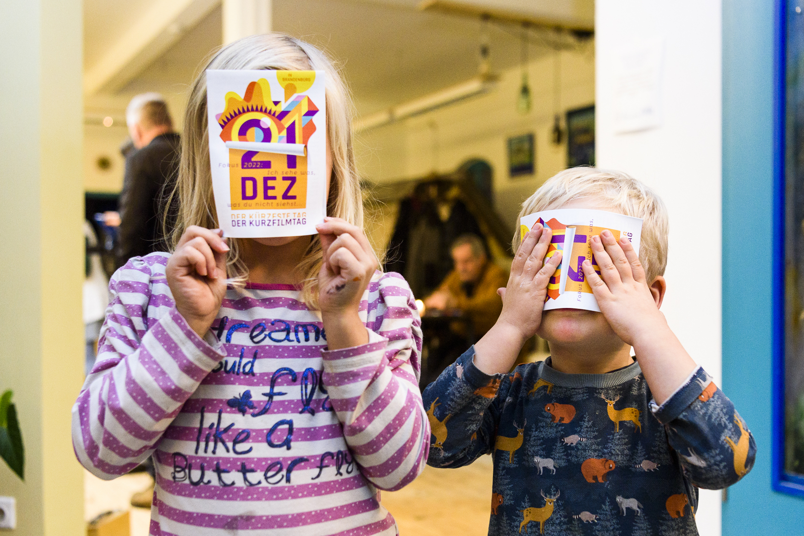 Zwei Kinder, die sich eine Postkarte mit dem Motiv des KURZFILMTAG 2022 vors Gesicht halten.